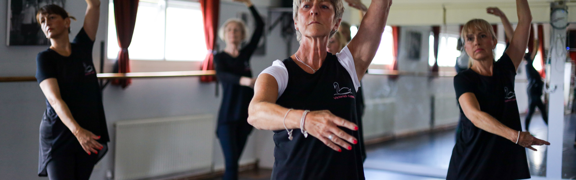 A group of four middle-aged women, all wearing black, holding a ballet pose, with one arm out in front of their bodies and one raised above their heads, with serious expressions on their face.