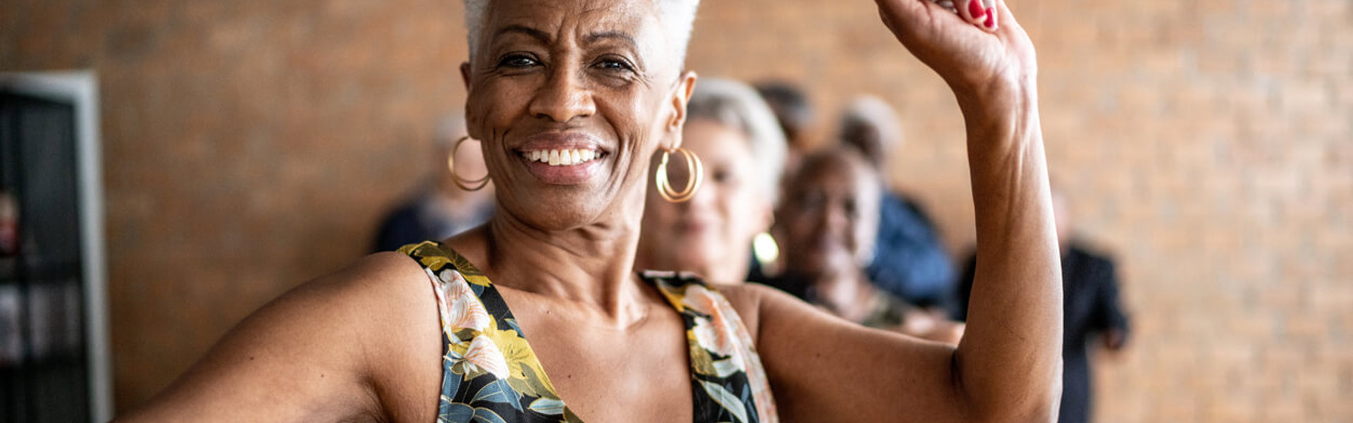 Woman dancing with one arm in the air, smiling wearing a black dress with a yellow and blue floral pattern.