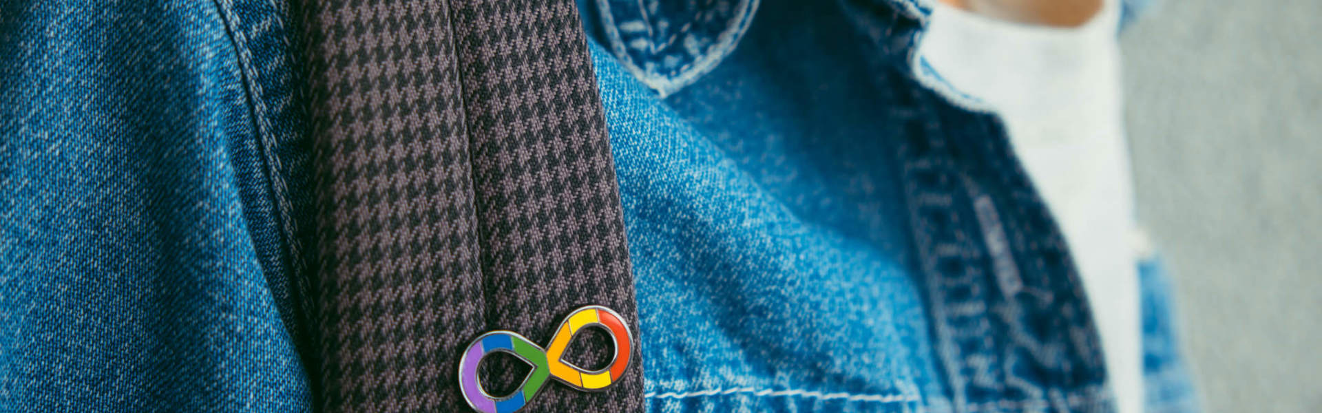 Person in a denim jacket, wearing a back pack and the rainbow infinity symbol badge, representing Autism Acceptance Month.